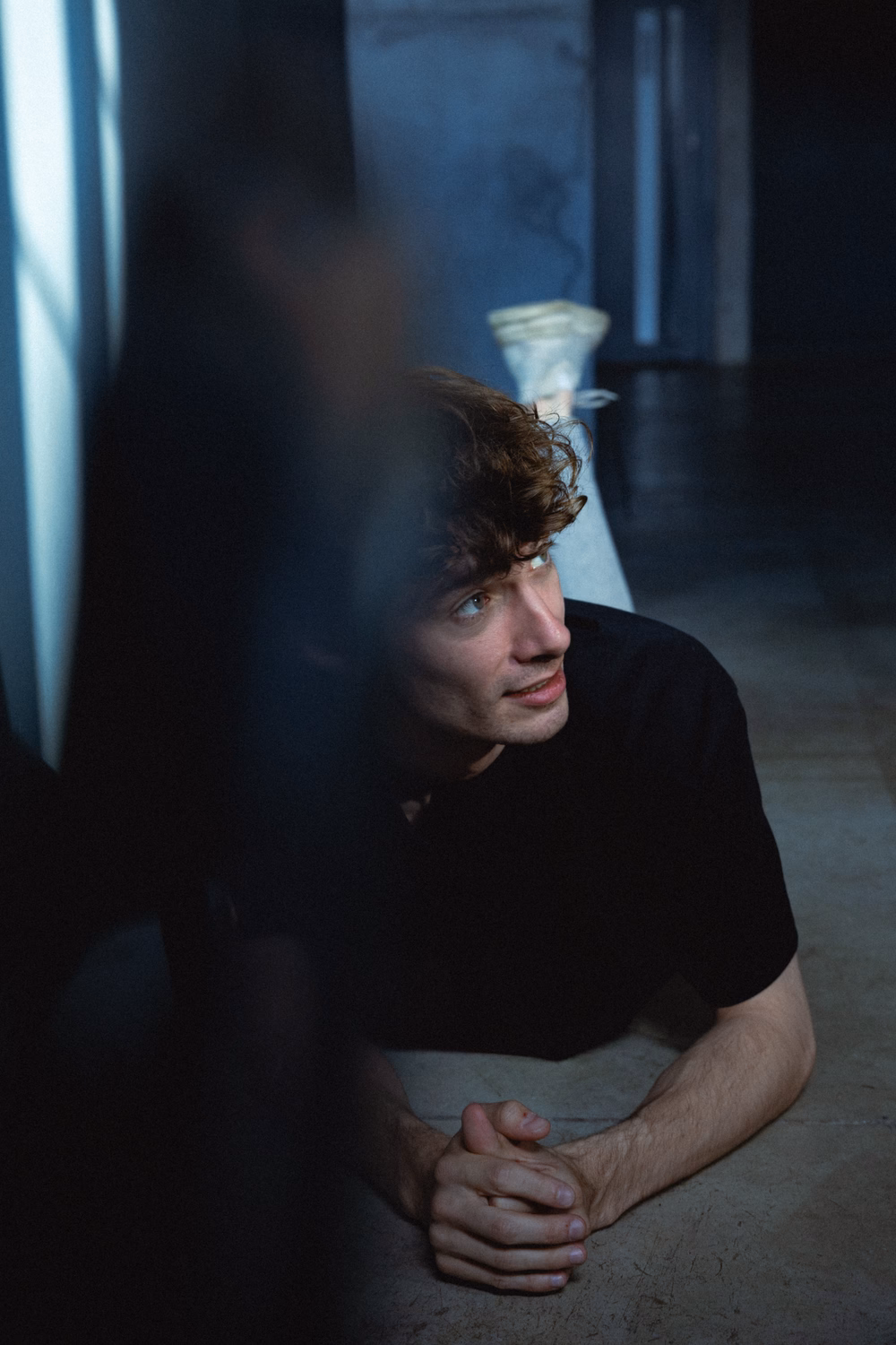 Ein Mann mit braunem Haar in schwarzem Shirt lehnt nachdenklich an einer dunklen Wand in schwachem Licht.