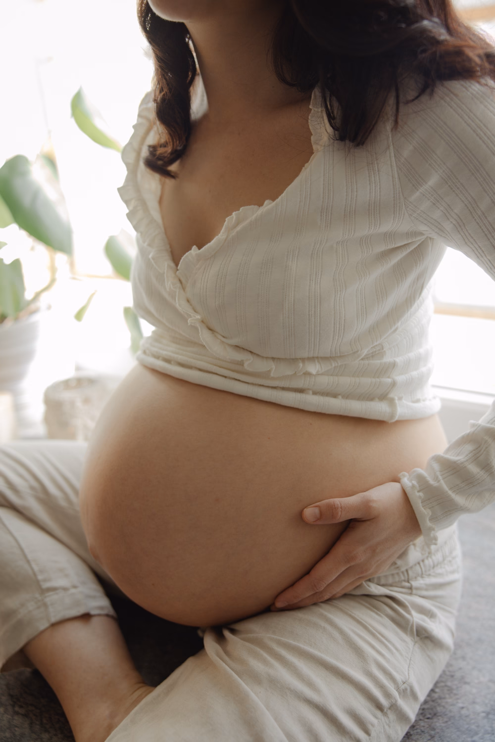 Schwangere Frau in weißem Oberteil hält ihren Bauch und sitzt neben einer Pflanze im Sonnenlicht.