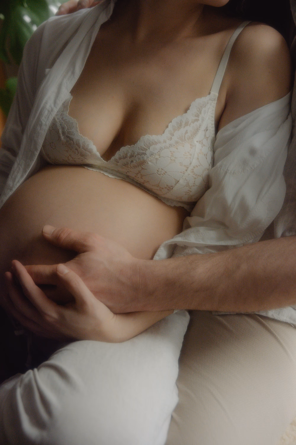 Schwangere Frau in weißer Wäsche, Partner umarmt sie zärtlich von hinten, Bauch berührend.