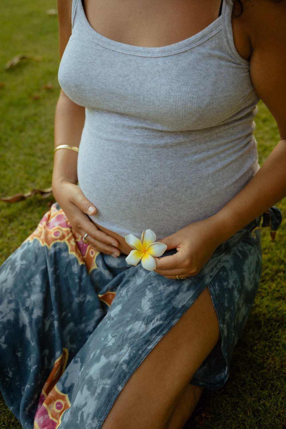 Schwangere Frau sitzt auf Gras und hält eine gelb-weiße Blüte in der Hand über ihrem Bauch.