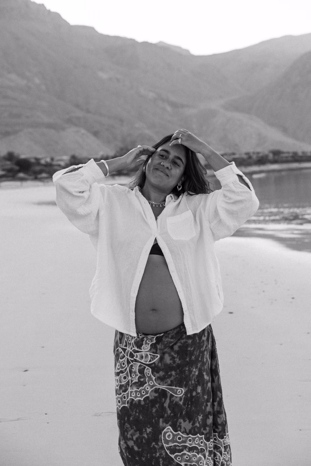 Schwangere Frau in weißem Hemd am Strand vor Bergen, Schwarz-Weiß-Fotografie.