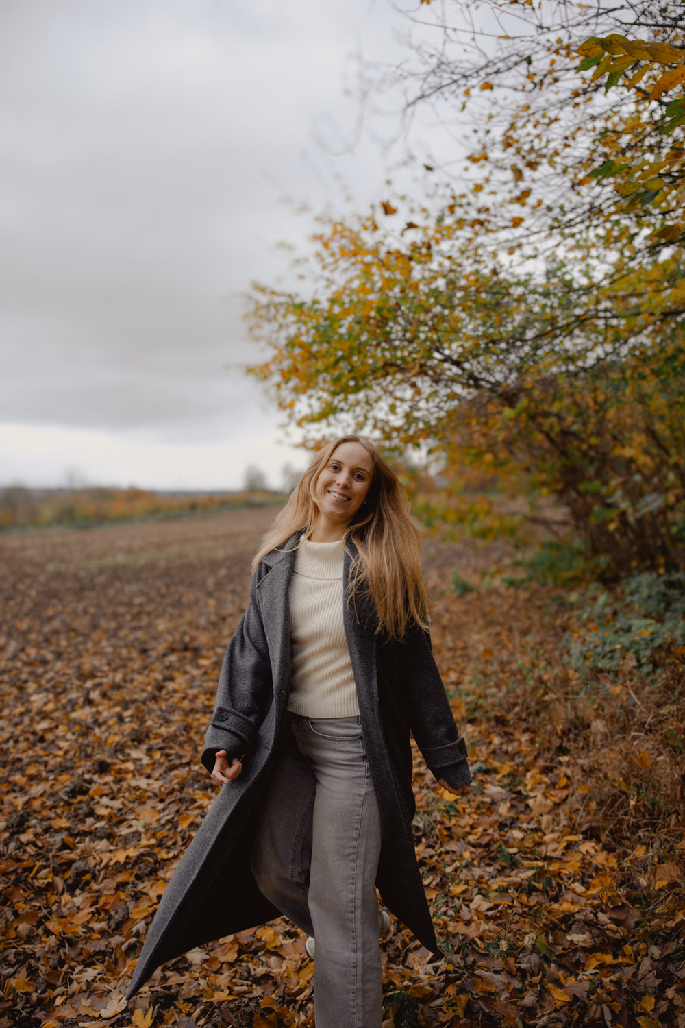 Junge Frau in grauem Mantel und weißem Pullover steht lächelnd auf einem Herbstfeld unter Bäumen mit gelben Blättern.