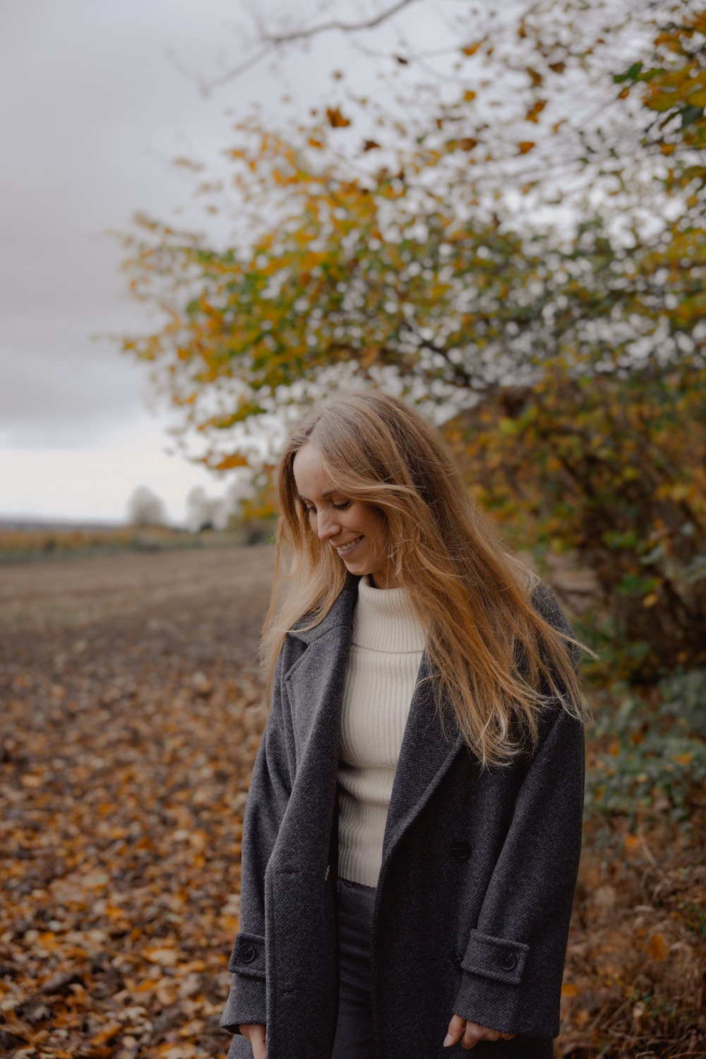 Junge Frau mit langen braunen Haaren in grauem Blazer und weißem Pullover unter blühendem Baum in Herbstlandschaft.