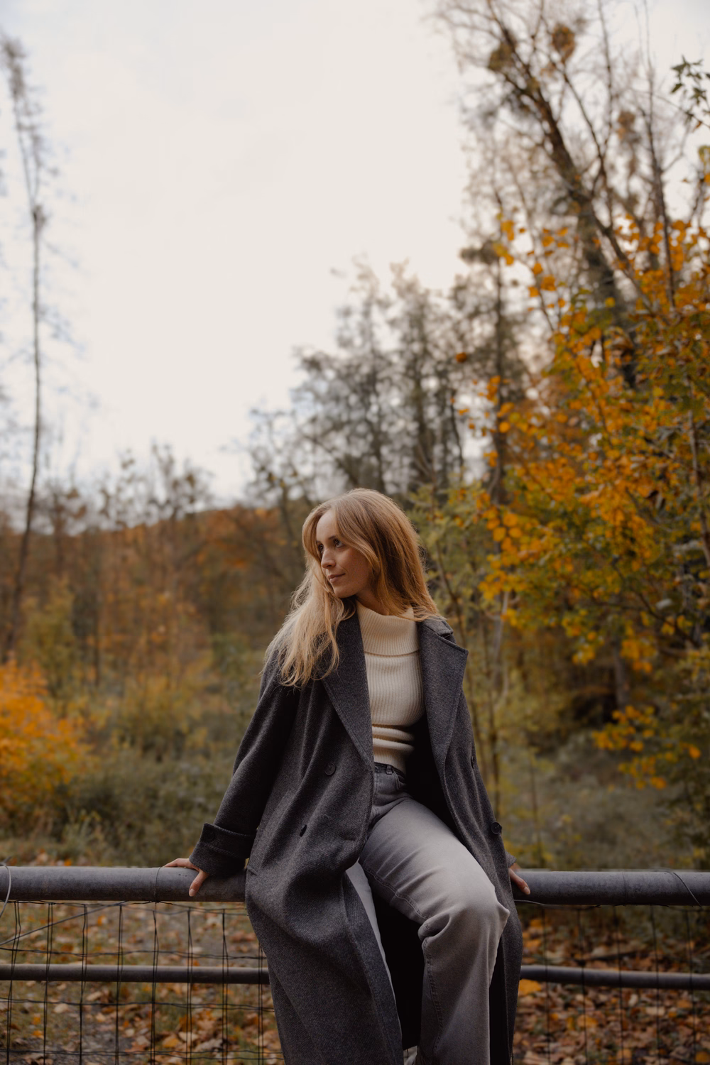 Junge Frau mit langen rötlichen Haaren sitzt auf einem Geländer in herbstlicher Waldlandschaft mit grauem Mantel.