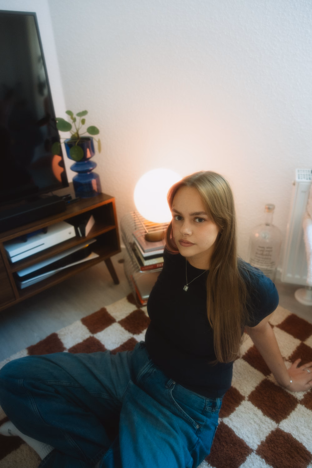 Junge Frau sitzt in Wohnzimmer, trägt schwarzes Shirt und Jeans, blickt zur Kamera, warme Beleuchtung.