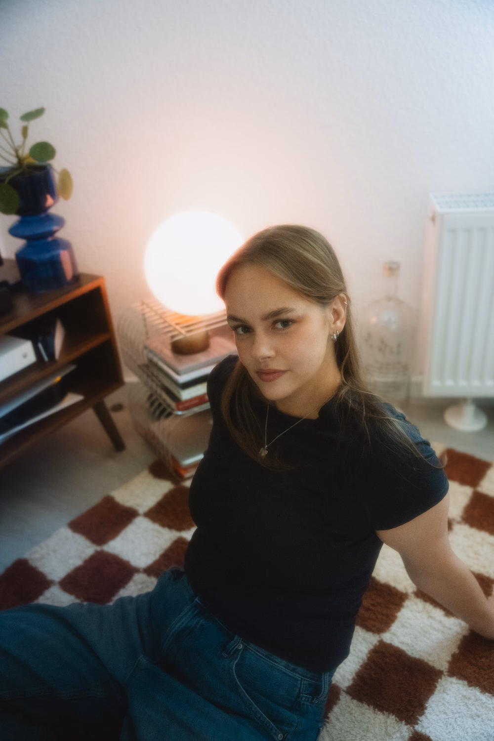 Junge Frau in schwarzem Shirt sitzt auf kariertem Teppich in gemütlichem Zimmer mit Lampe und Pflanze.
