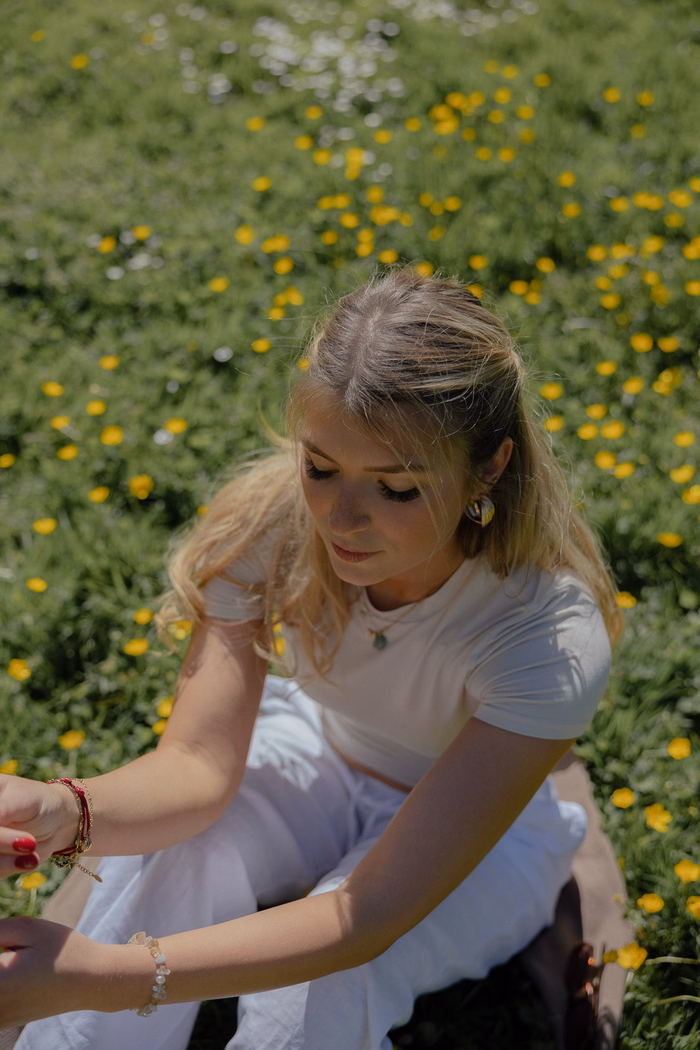 Junge Frau in weißem T-Shirt sitzt in einem Feld mit gelben Blüten und lächelt nach unten.