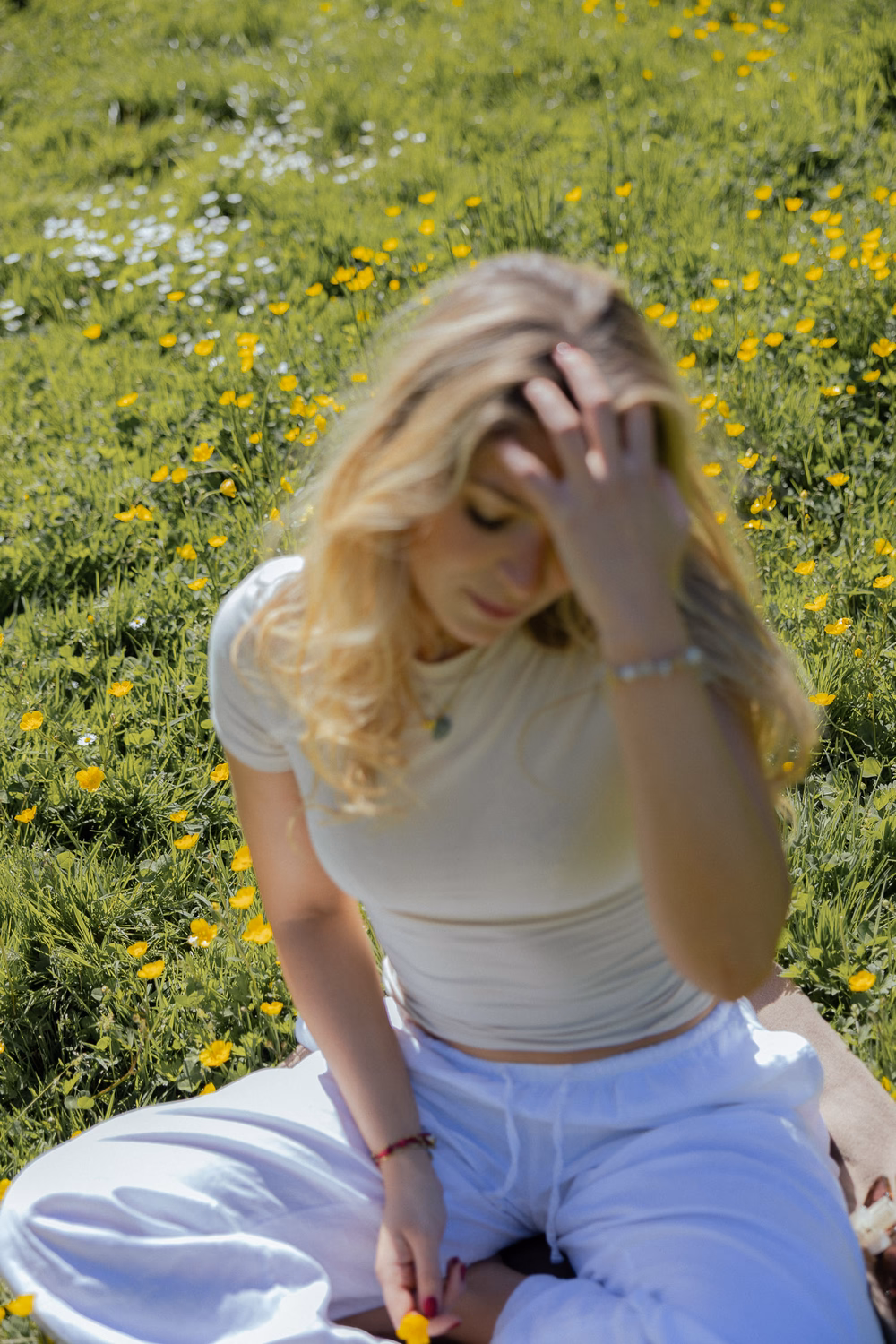 Junge Frau sitzt in Blumenwiese mit gelben und weißen Blüten, trägt graues Top und blaue Hose, Hand im Haar.
