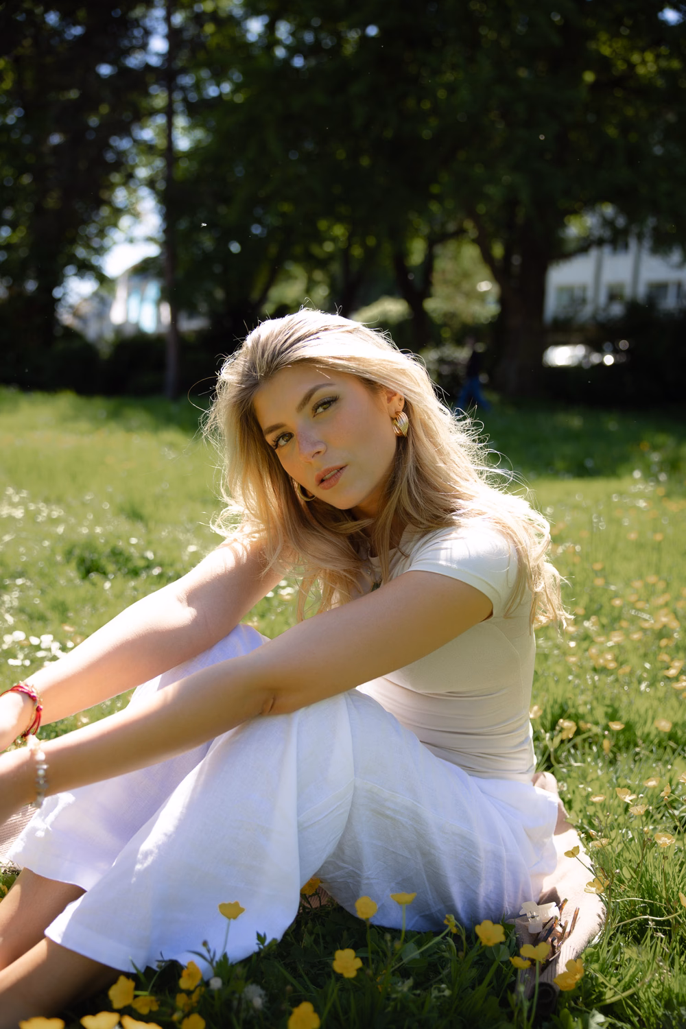 Blonde Frau sitzt auf grüner Wiese mit gelben Blüten, trägt weißes Oberteil und Rock, sonniger Park im Hintergrund.