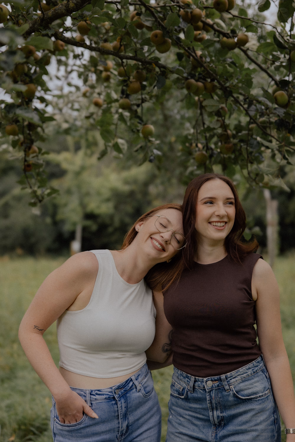 Zwei junge Frauen lachen zusammen unter einem Obstbaum mit grünen Früchten in einem Park.