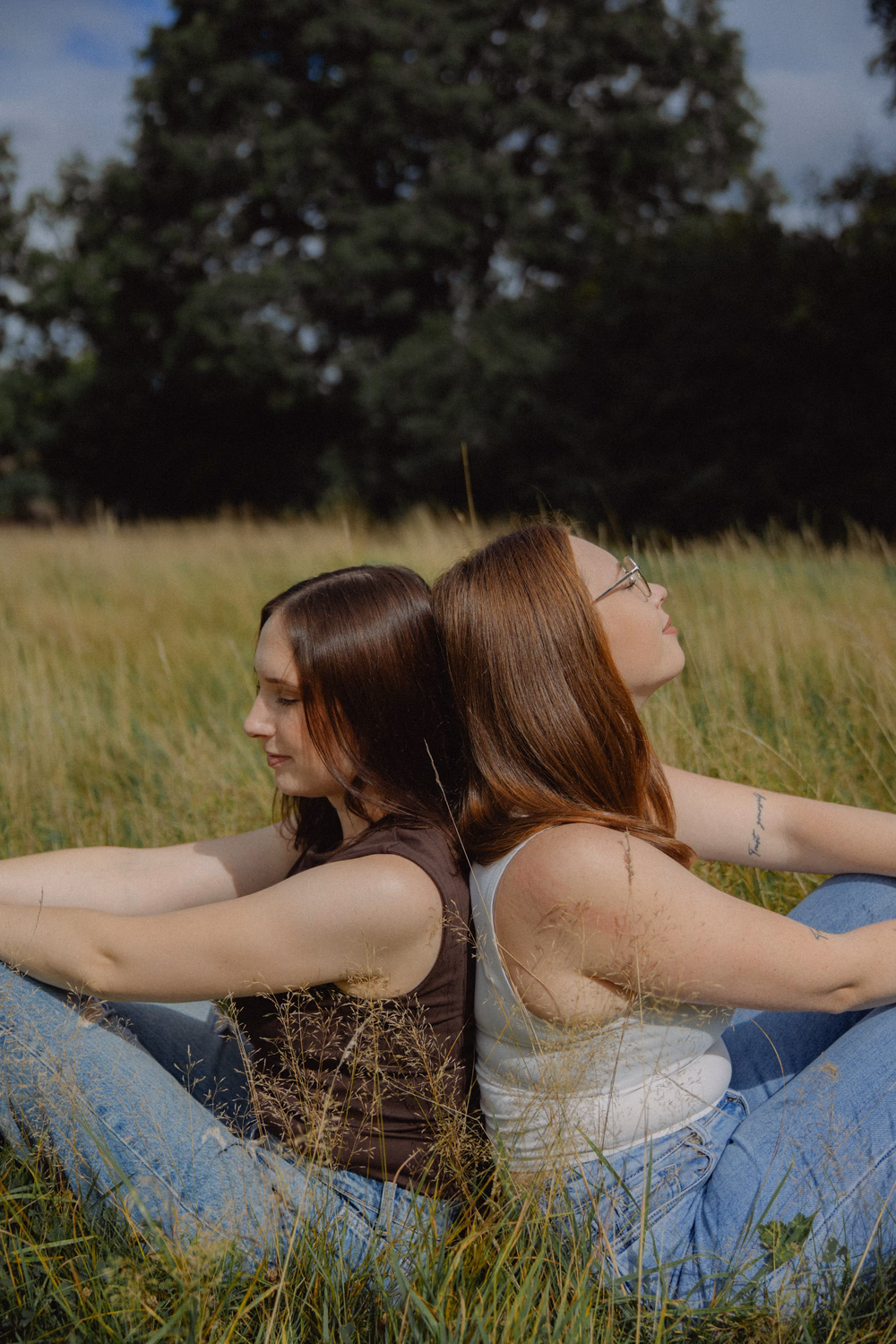 Zwei junge Frauen sitzen Rücken an Rücken in einem Grasfeld mit Wald im Hintergrund und blicken zur Seite.