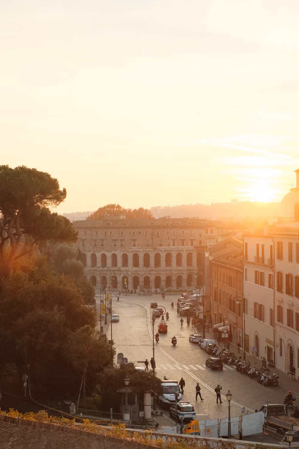 Sonnenaufgang über einem italienischen Platz mit historischen Gebäuden, Bäumen und Fußgängern bei goldenem Morgenlicht.