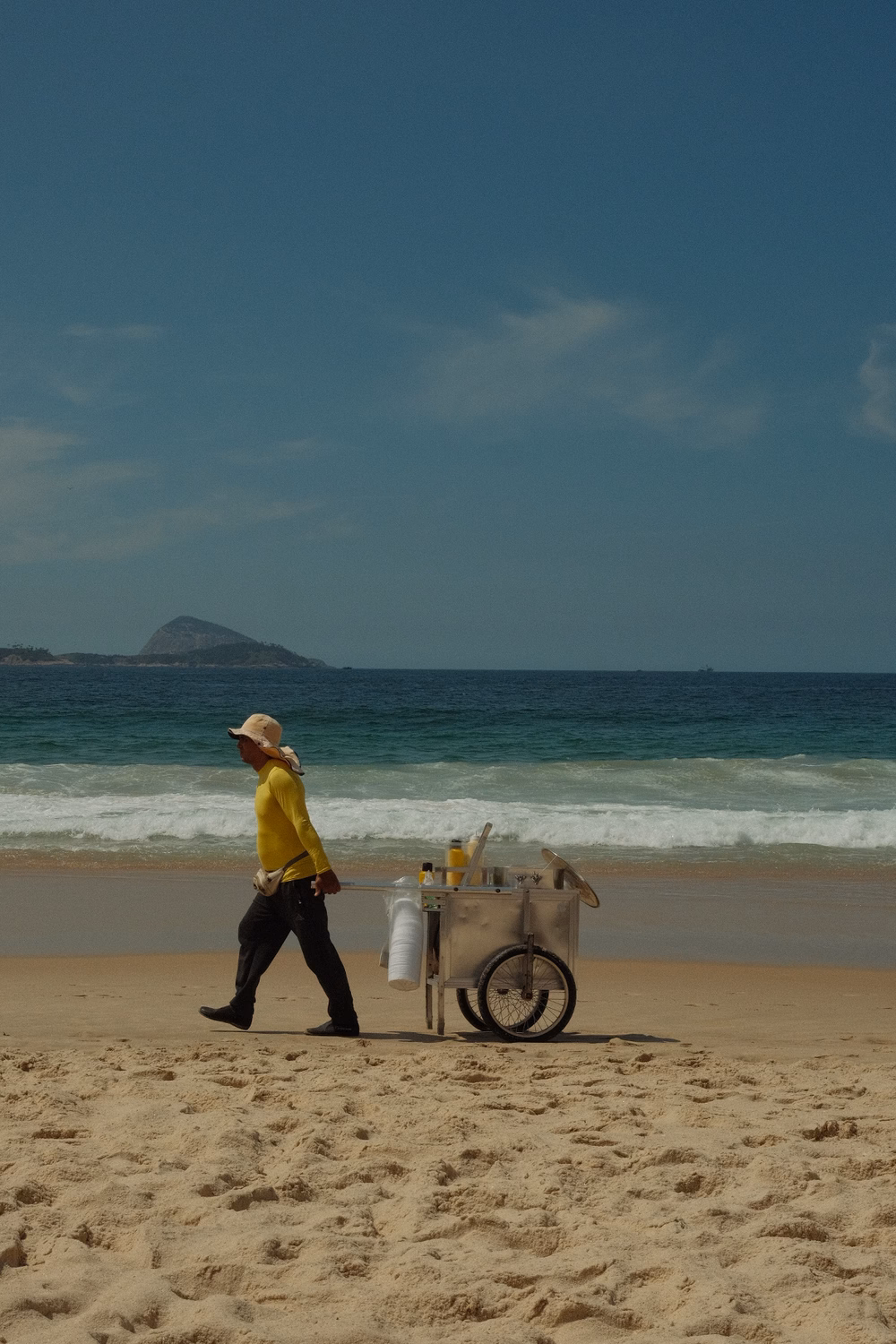 Straßenverkäufer mit gelbem Hemd schiebt Verkaufswagen am Strand entlang, Meer und Insel im Hintergrund.