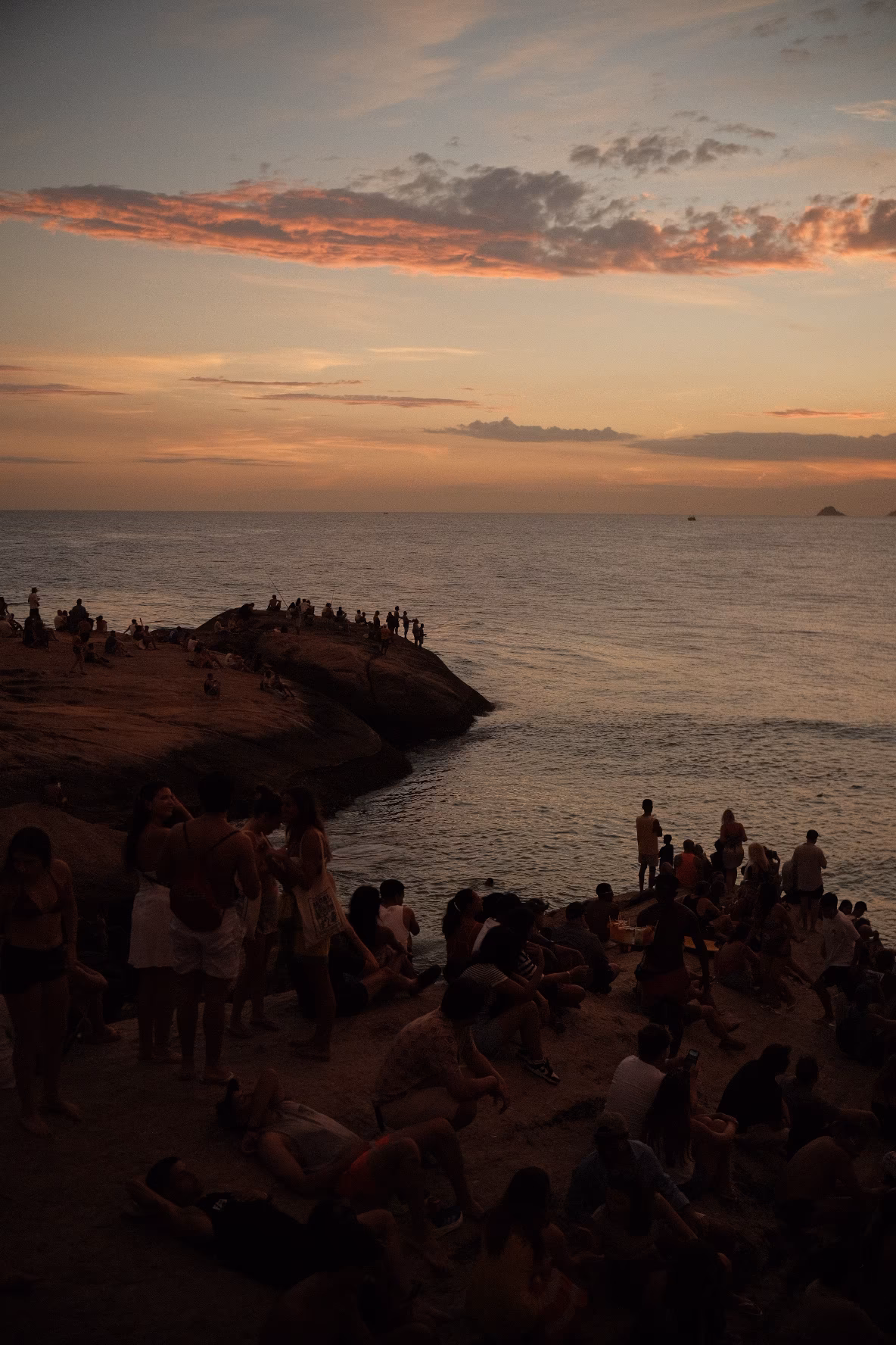Strand bei Sonnenuntergang mit Besuchern, Felsen und ruhigem Meer unter dramatischem Himmel.