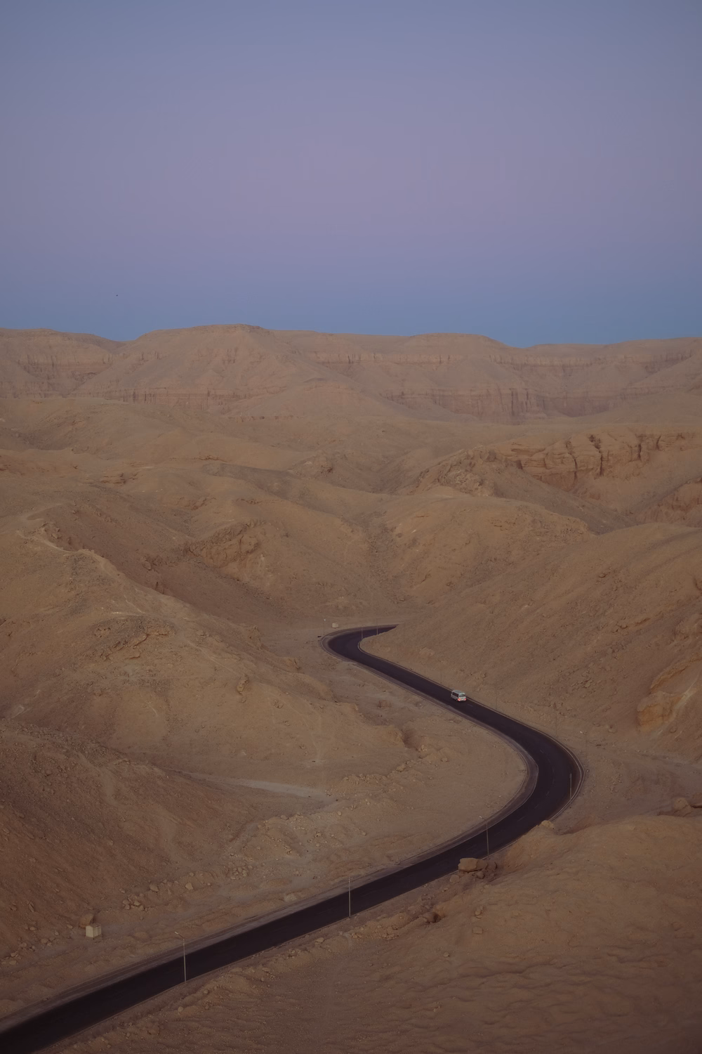 Asphaltstraße schlängelt sich durch karge Wüstenlandschaft mit sandfarbenen Hügeln und klarem blauen Himmel.