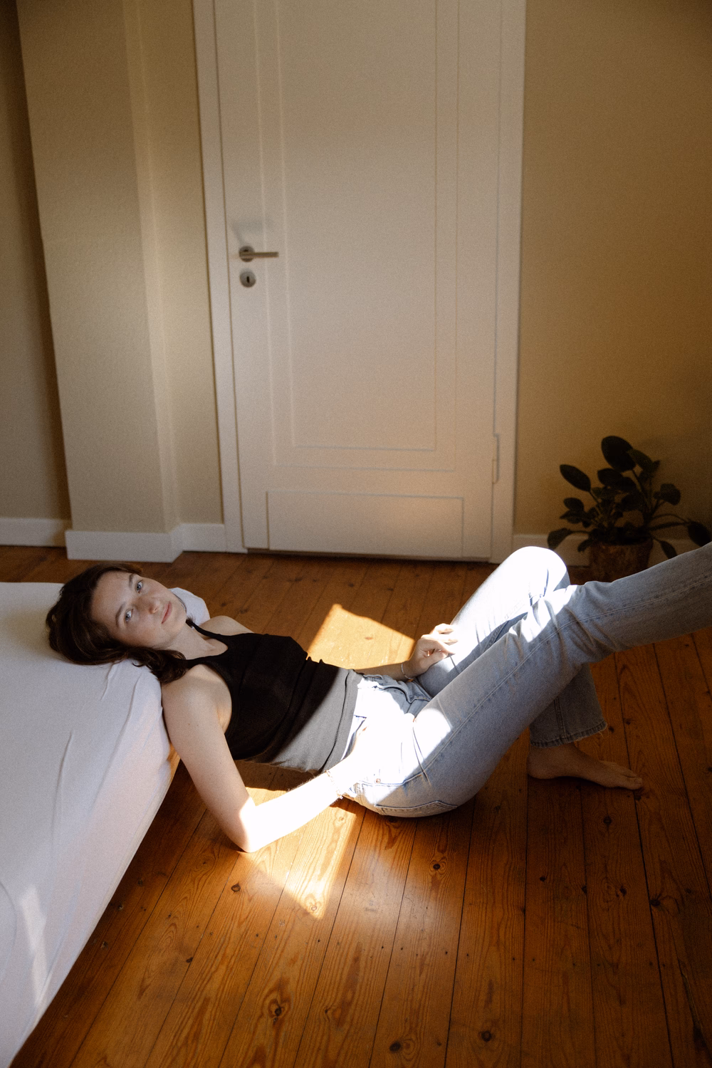 Frau liegt auf Holzboden in Sonnenlicht, trägt schwarzes Top und Jeans, liest Buch in modernem Zimmer.