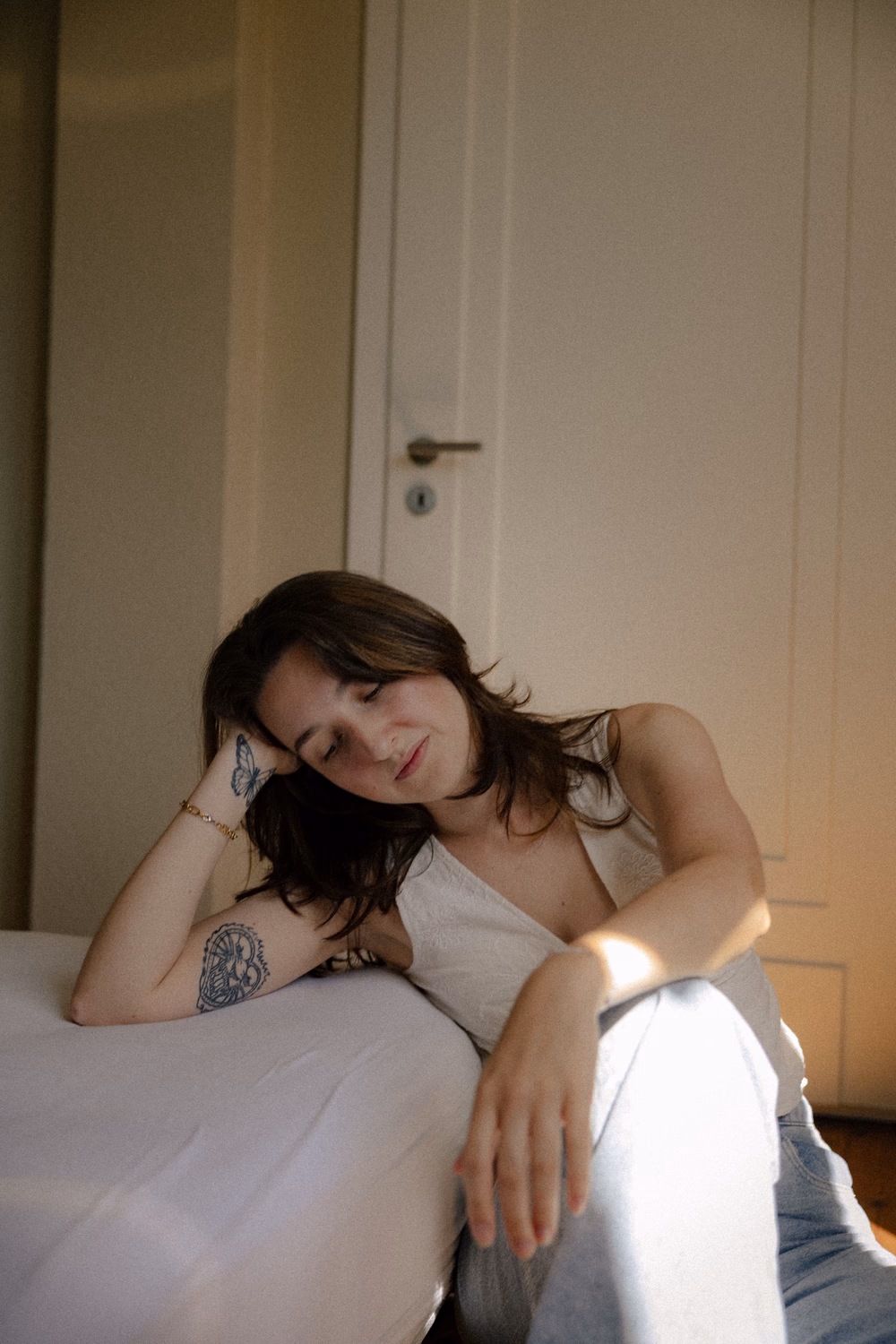 Frau mit Tattoos sitzt auf Bett, trägt weißes Top, sonniges Licht im minimalistischen Raum.