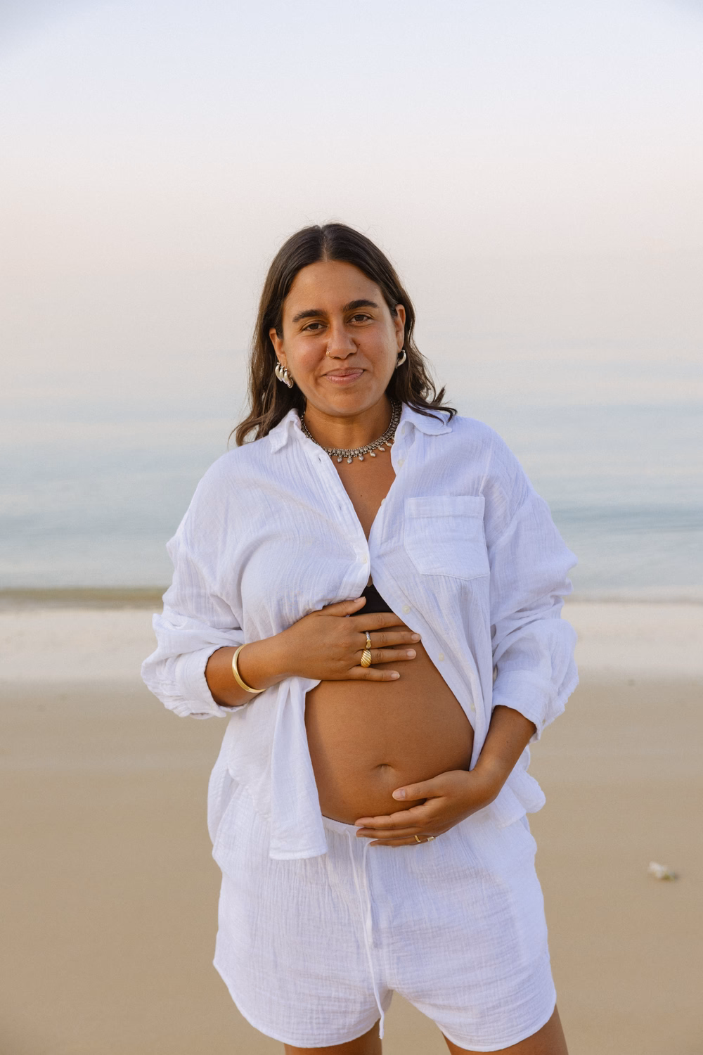 Schwangere Frau in weißem Hemd am Strand, hält ihren Bauch und lächelt zur Kamera.