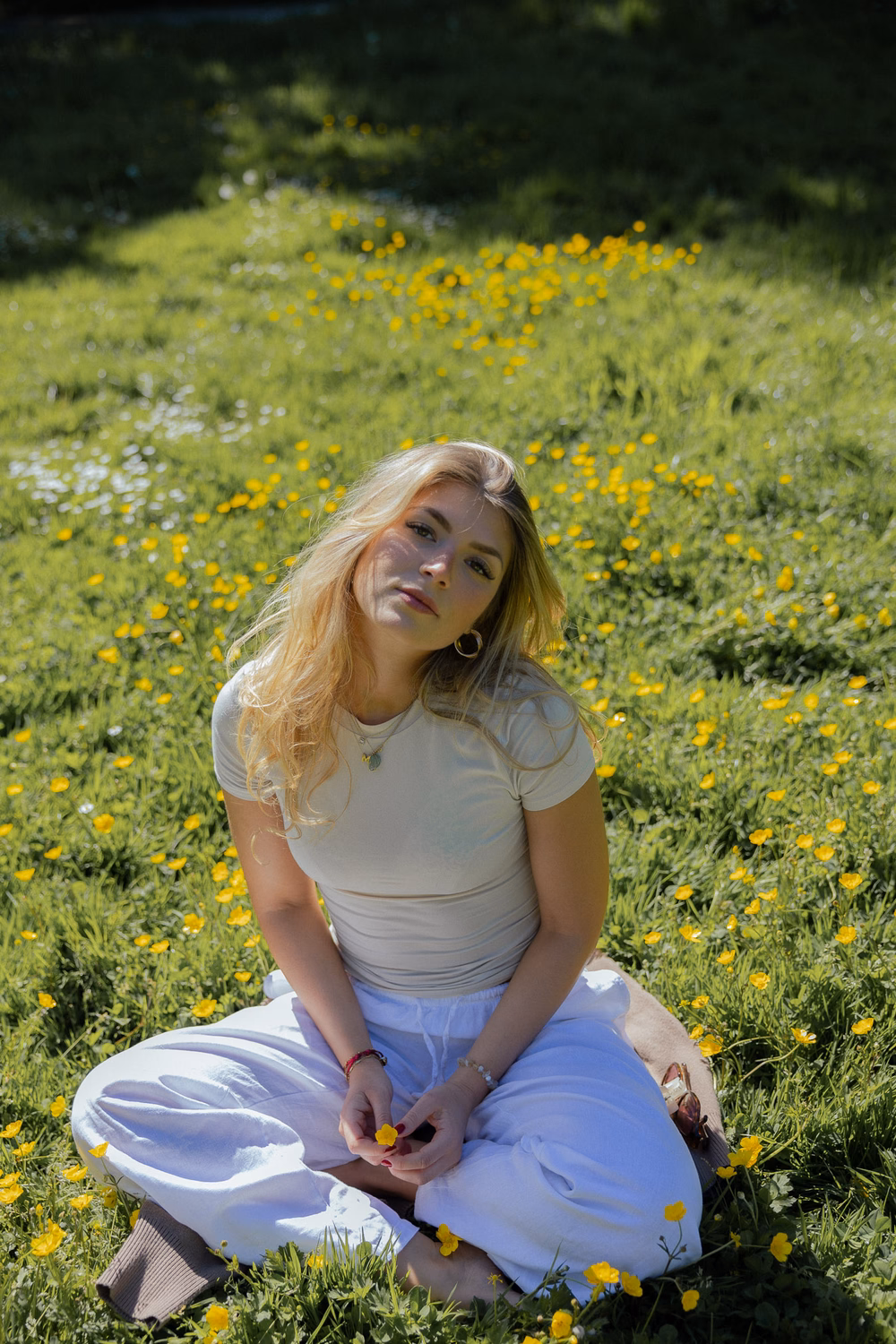 Junge Frau sitzt in einem Blumenfeld mit gelben Blüten, trägt weißes Shirt und blaue Hose, schaut nach oben.