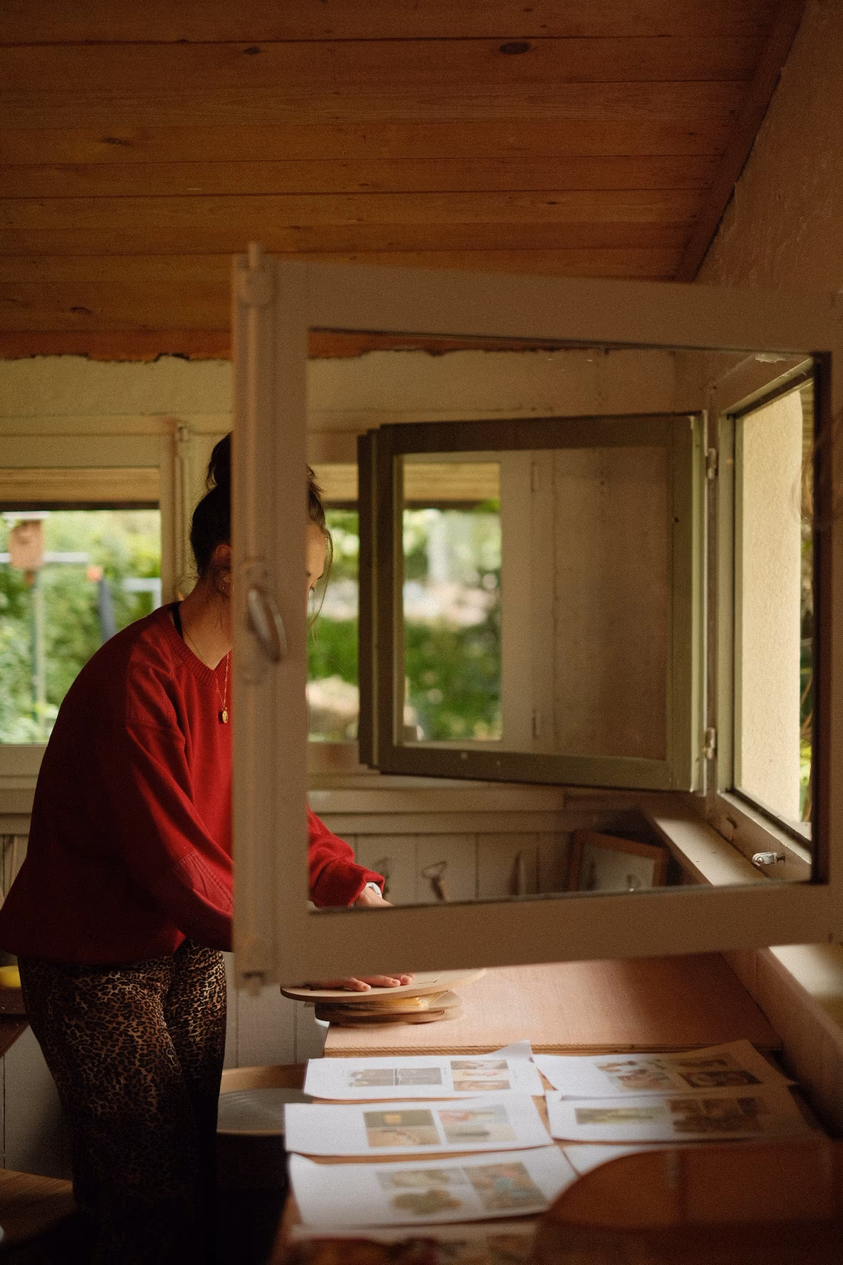 Frau in rotem Pullover schaut durch offenes Fenster, Papiere auf Tisch im Vordergrund, Holzdecke.