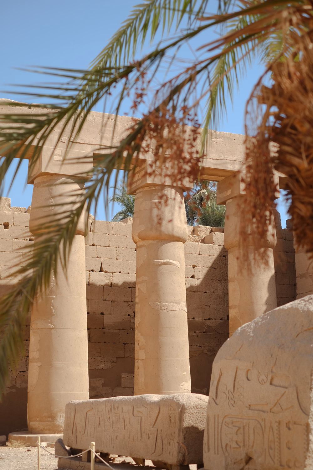 Antike ägyptische Tempelruinen mit Säulen, Hieroglyphen und Palmenblättern unter blauem Himmel.