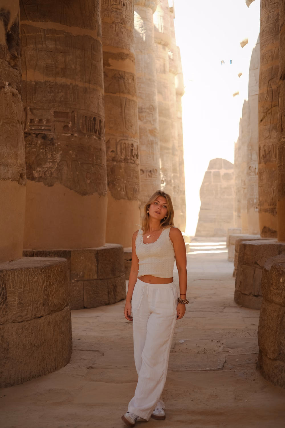 Frau in weißem Outfit zwischen antiken ägyptischen Säulen mit Hieroglyphen in staubiger Atmosphäre.