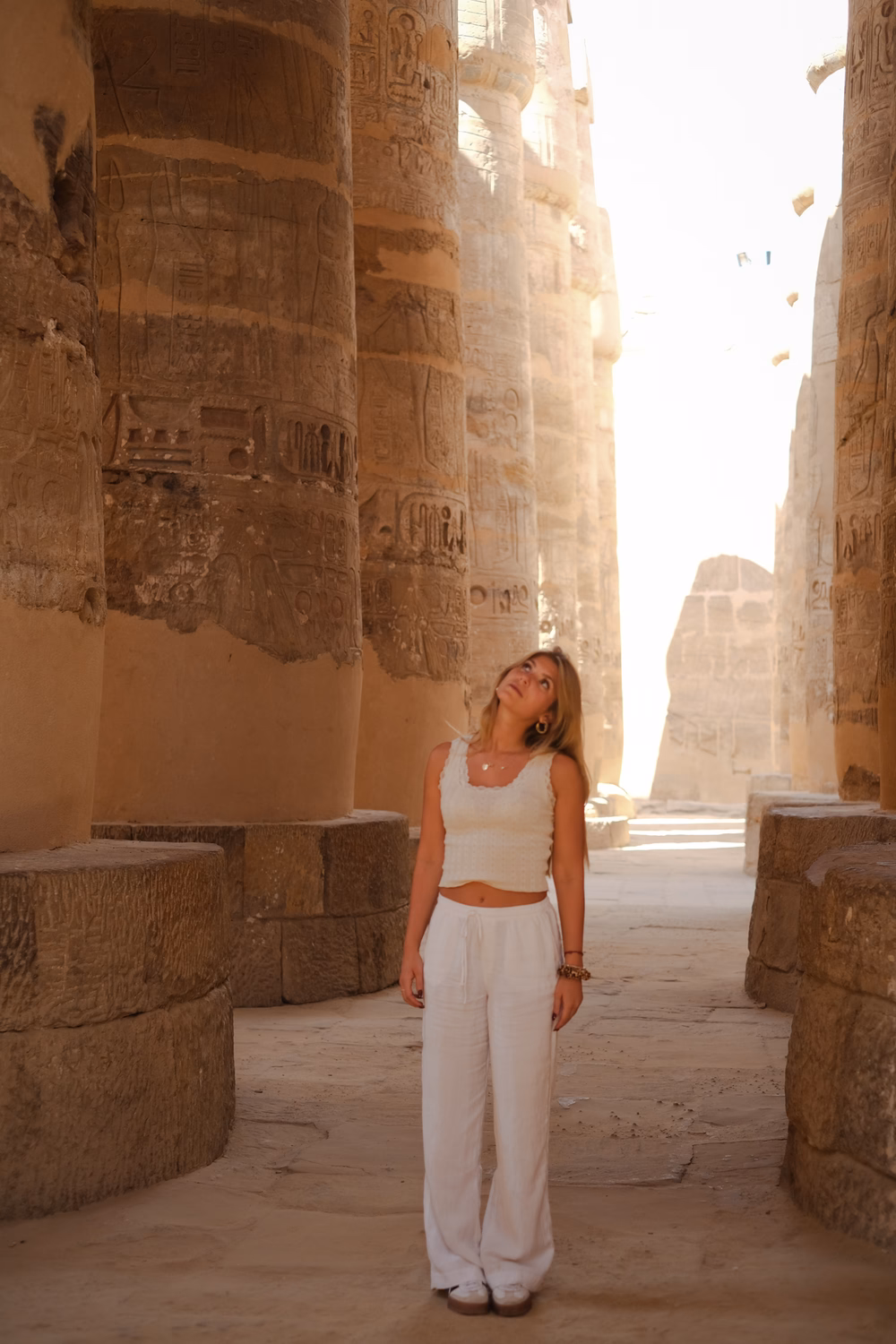 Frau in weißem Outfit steht zwischen massiven Säulen mit ägyptischen Hieroglyphen in einem antiken Tempel.