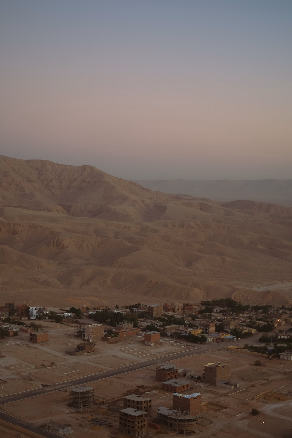Wüstensiedlung mit Lehmhäusern unter kahlen, sandfarbenen Bergen in Ägypten oder Nordafrika bei Dämmerung.