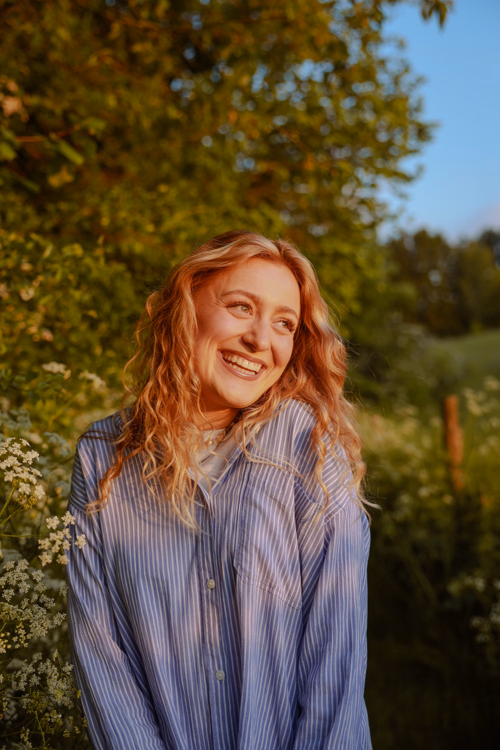 Junge Frau mit roten Locken lächelt in einem Garten zwischen weißen Blüten und grünem Laub in der Sonne.