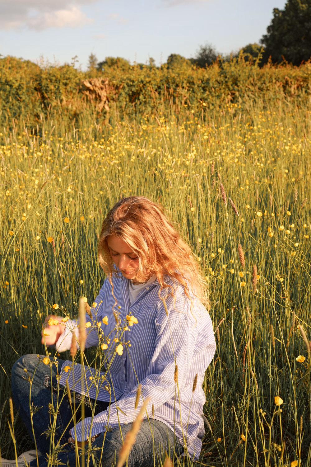 Frau mit rötlichem Haar sitzt in einem Wildblumenfeld mit gelben Blüten und grünen Gräsern.