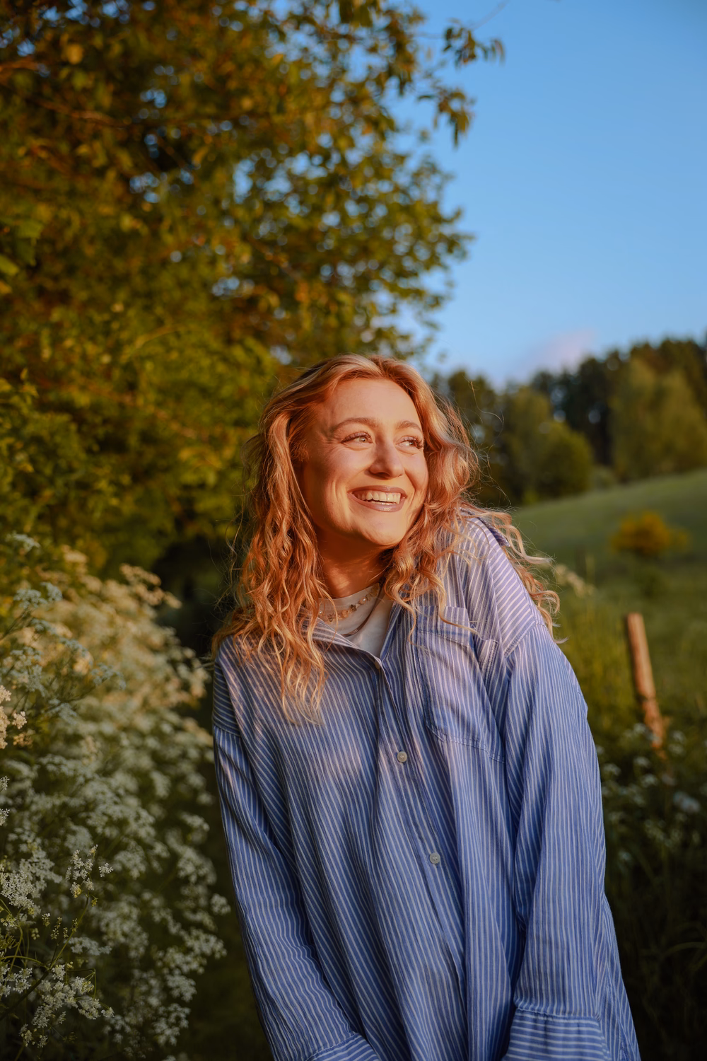 Junge Frau mit rötlichen Haaren lächelt in einer grünen Landschaft mit Bäumen und Blüten.