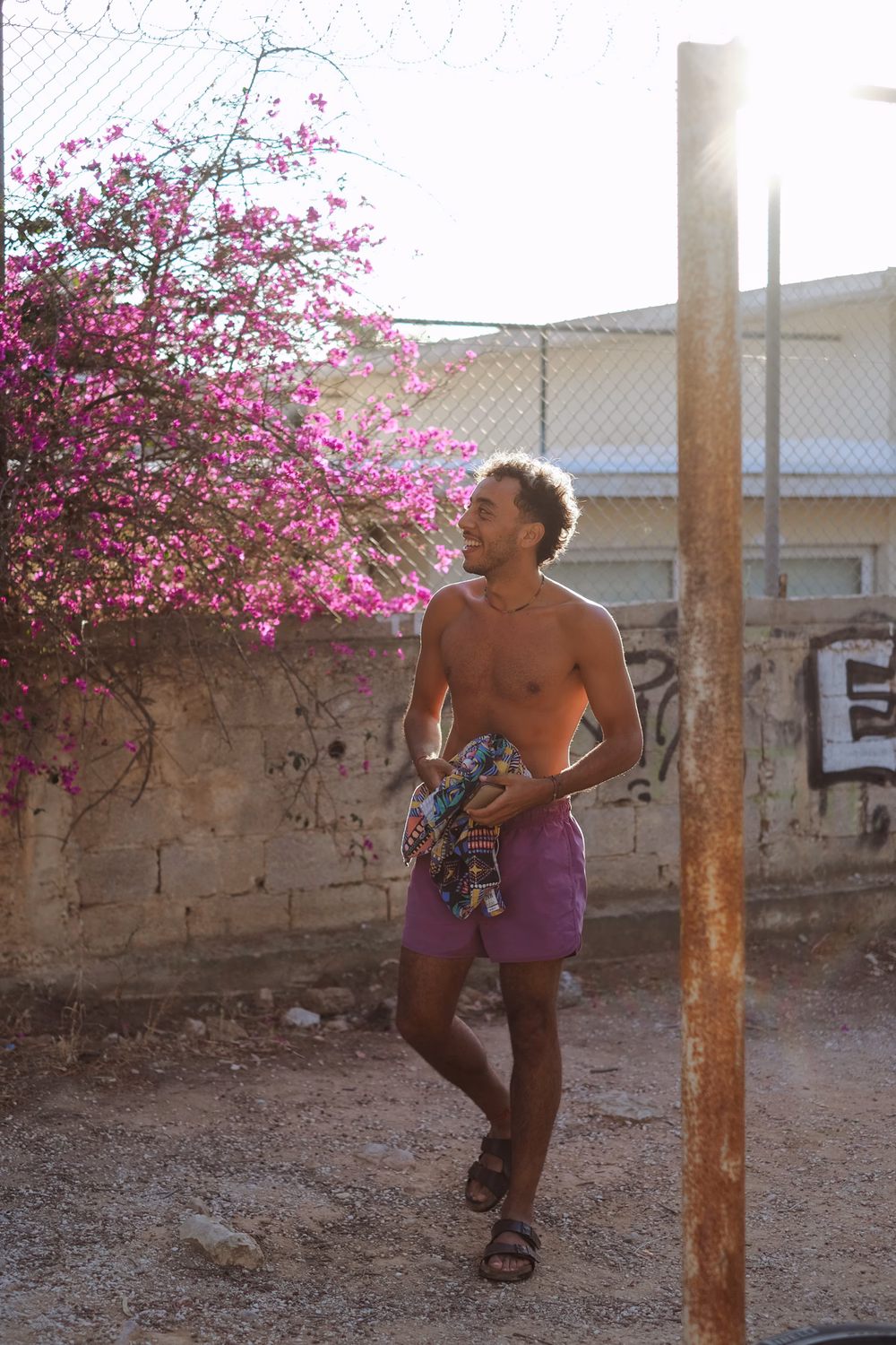 Ein lächelnder Mann in lila Shorts steht neben einer blühenden Magentablüte in einem ummauerten Hof.