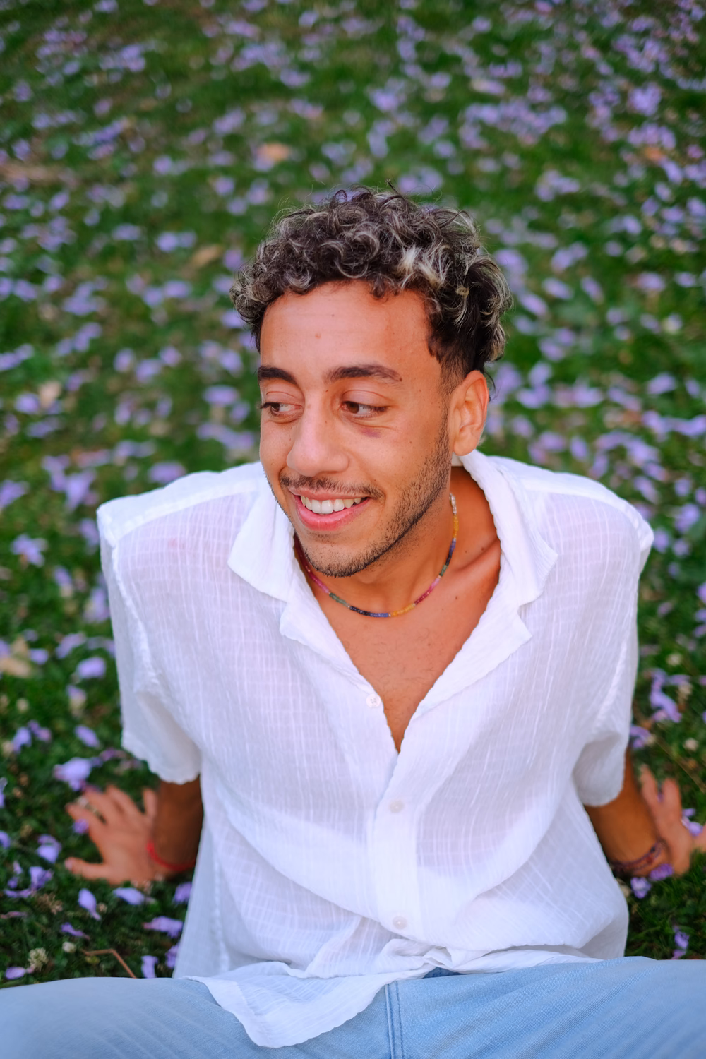 Ein lächelnder Mann mit lockigem dunklem Haar sitzt vor blühenden lila Blumen in einem weißen Hemd.
