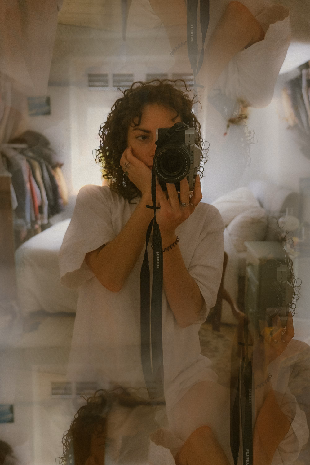Frau mit lockigem dunklem Haar macht Spiegelselfie mit analoger Kamera im hellen Zimmer.